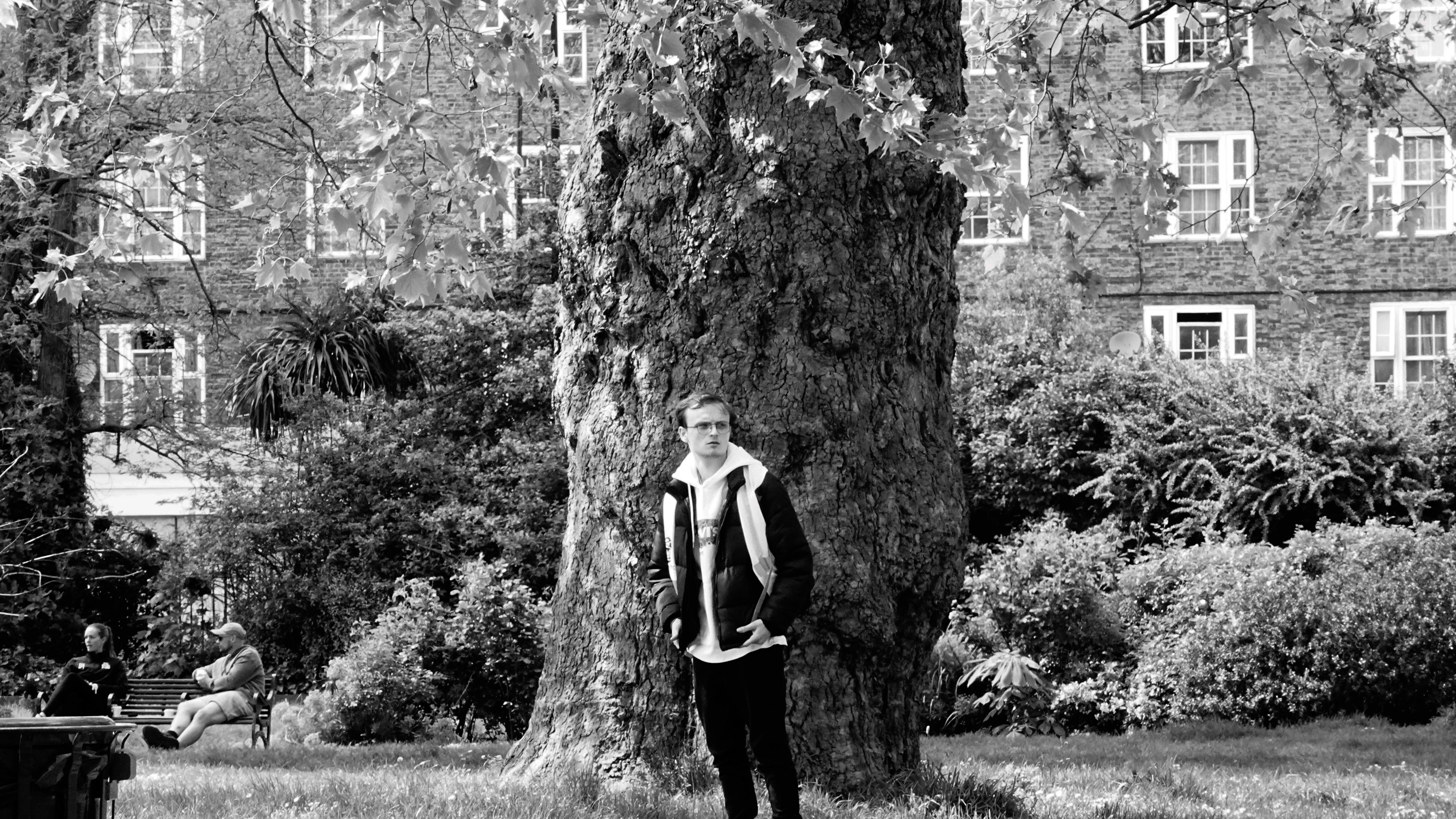 A young man in a park stands in front of a tree, his body facing one way while he looks in another.