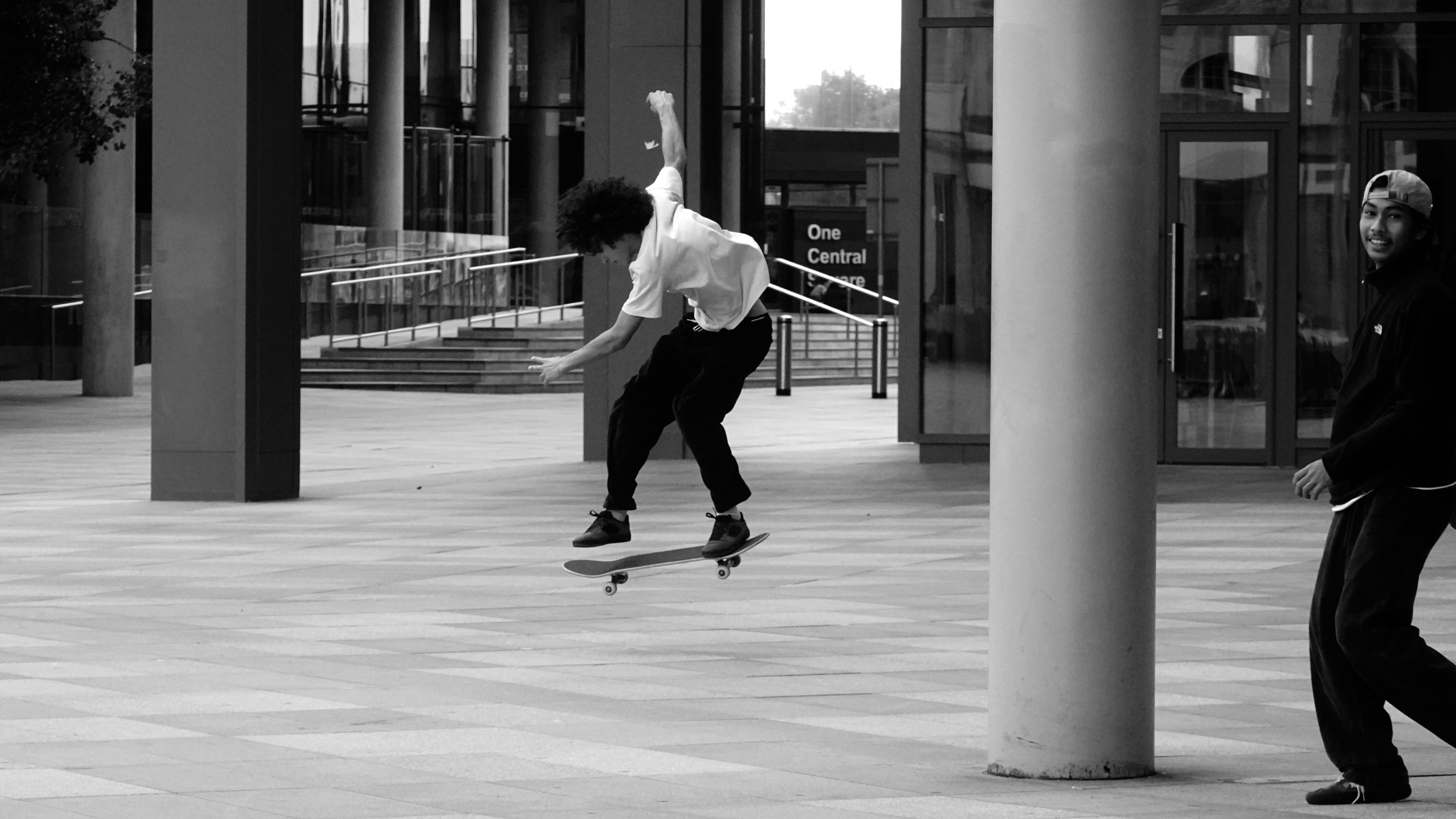 A black and white image of a skateboarder, returning to the ground after a trick.  His friend smiles at the camera.